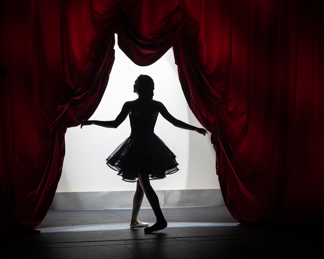 A silhouette of a female ballet dancer (Rylee Rogers) stands gracefully between two large, parted red stage curtains. She wears a dark, knee-length tutu and ballet slippers. Her arms are extended gently to either side, holding the curtains open. Bright white light from the stage or backstage area behind her outlines her form, emphasizing her poised stance. The scene conveys a moment of anticipation or transition, as if she is about to enter or exit the stage.