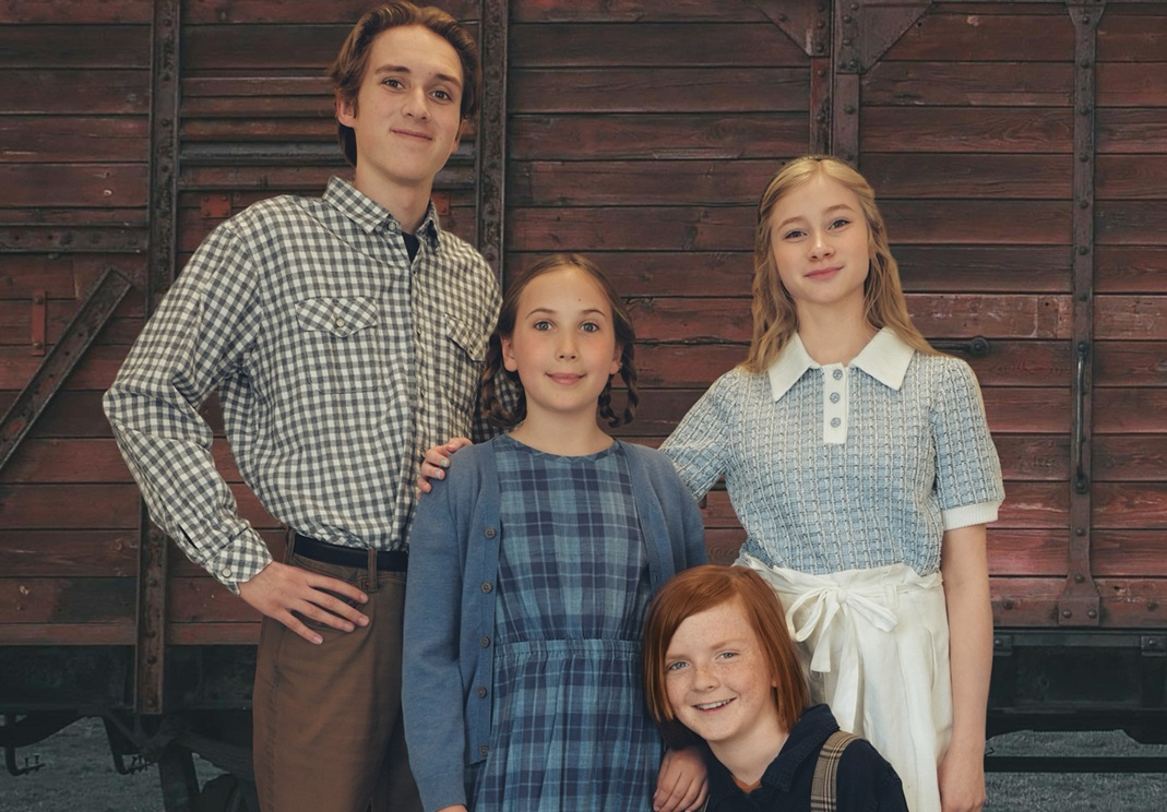 A promotional banner features four young actors in period costumes standing in front of a weathered red wooden boxcar. From left to right: a tall teenage boy in a checkered button-down shirt and brown pants stands with his hand on the shoulder of a younger girl in a blue plaid dress and cardigan. Next to her, a blond teenage girl in a short-sleeved textured knit top and cream skirt smiles warmly. Kneeling in front is a red-haired boy in navy overalls and a dark shirt, also smiling. Text below the group reads: “The Box-car Children by Melissa Leilani Larson – Creekside Theatre Fest 2025.”