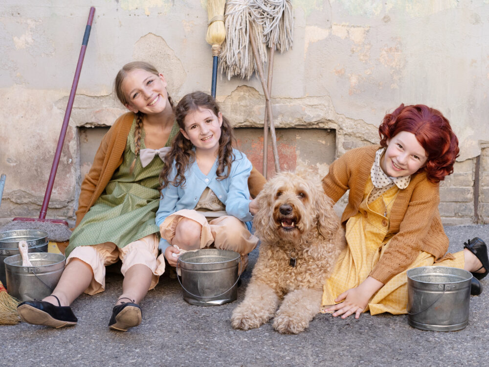 Three young girls dressed as orphans in vintage-style costumes sit on the ground outside against a weathered wall, smiling brightly. Each wears a cardigan over a simple dress with worn shoes and has props like metal cleaning buckets nearby. Between them lies a fluffy, golden-colored dog (playing Sandy) looking directly at the camera with its tongue slightly out. Behind them are cleaning supplies including a mop and broom, enhancing the scene’s setting as part of a musical theater production of Annie.