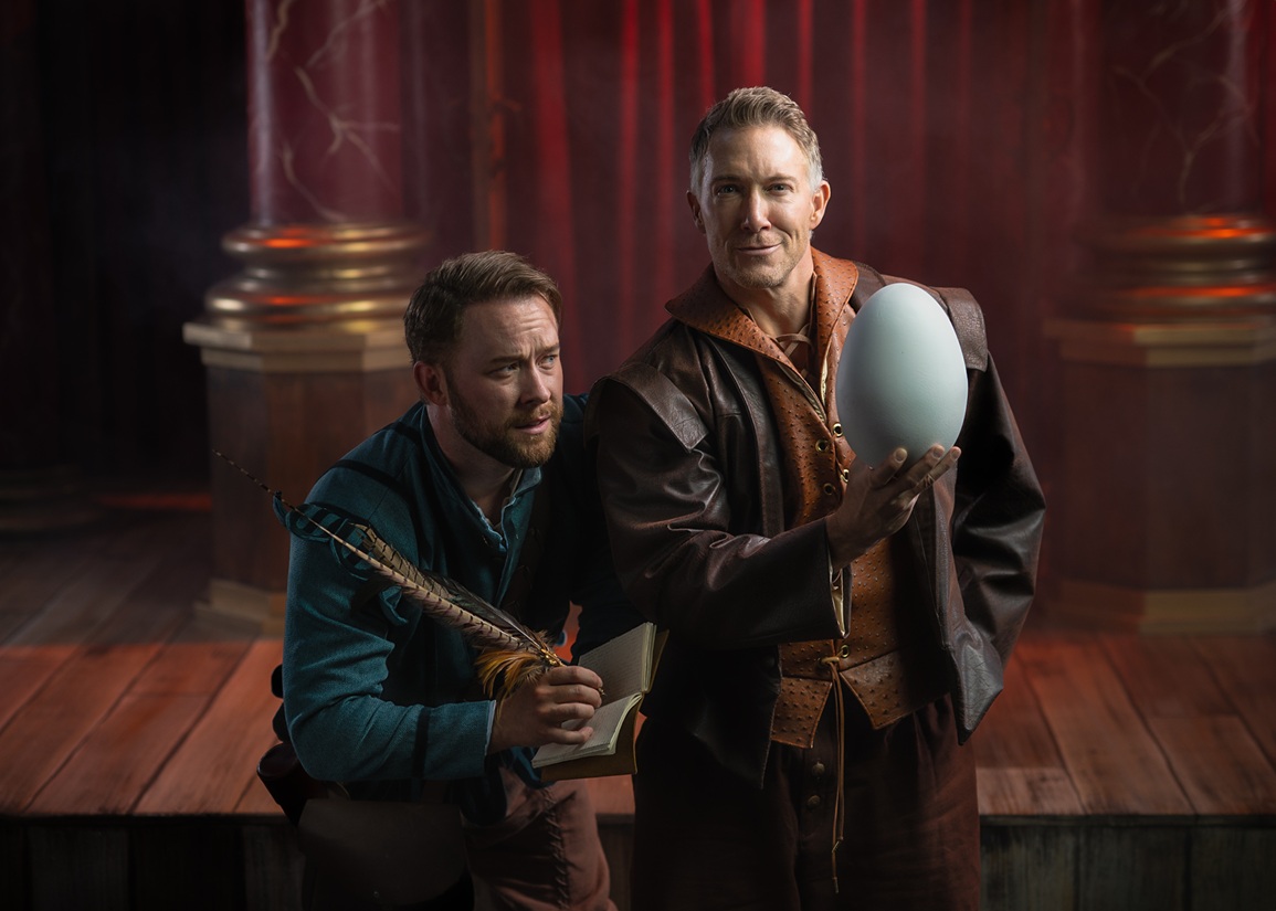 The image features two men on a stage with a theatrical backdrop. The man on the left is kneeling and gazing thoughtfully upward. He is dressed in a teal tunic and holds a quill and an open book. The man on the right stands confidently, wearing a brown leather coat over an orange vest. He holds a large, white egg-shaped object in his right hand. The background includes rich red curtains and decorative columns, creating a dramatic setting.