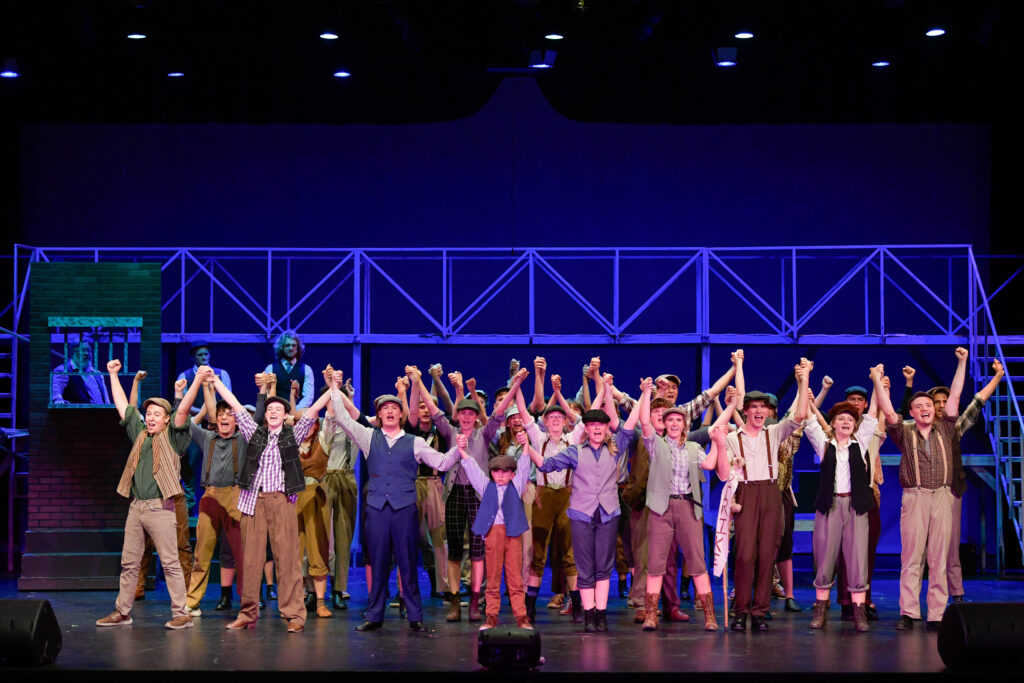 The image shows a scene from the musical "Newsies" with a large group of performers on stage. They are standing in a horizontal line, holding hands and raising them triumphantly. The performers are dressed in period costumes typical of early 20th-century newsboys, including vests, plaid shirts, suspenders, and flat caps. The stage is lit with bright blue and purple lighting, creating a vibrant atmosphere. In the background, there is a structure with a scaffold-like design, suggesting an urban setting. A dark brick wall is visible to the left with three people inside what seems to be a window, watching the performance. The overall composition conveys a sense of unity and celebration.