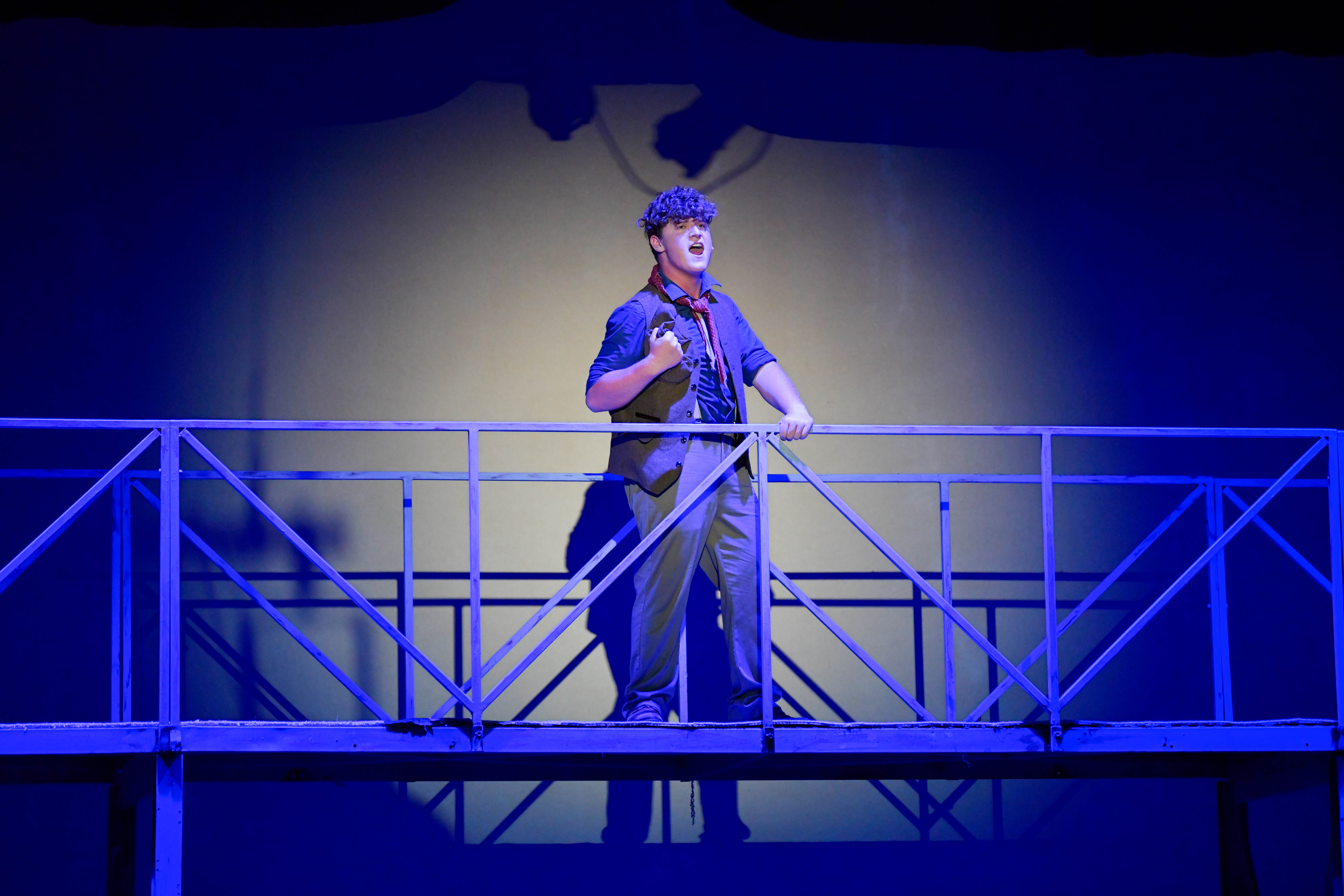 The image depicts a stage scene featuring an actor standing on a metal bridge structure. The background is a plain wall lit with a warm light creating a shadow behind the actor. The lighting casts a strong blue tint across the entire scene, adding drama and mood. The actor, positioned centrally, is wearing a dark shirt with a vest, trousers, and a red neckerchief. His posture is expressive, with one hand gripping the railing and the other placed near his chest, suggesting he is mid-performance or singing. The bridge's framework is geometric, consisting of straight lines and angles.