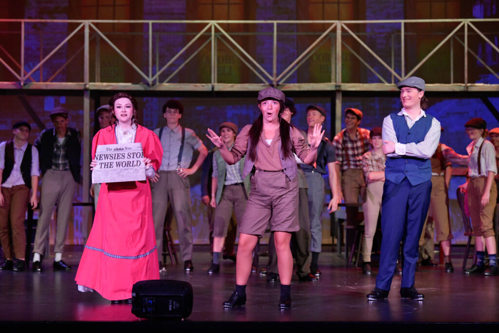 The image depicts a lively stage performance featuring a group of actors in costume, suggesting a theatrical production. At the center, a woman in a bright red period-style dress holds up a newspaper headline. Next to her, another performer dressed in early 20th-century men's attire, including a cap, appears to be energetically gesturing with hands raised. To their right, a man in a blue waistcoat and cap stands with folded arms, smiling. In the background, several other performers in similar vintage outfits appear, most of them wearing caps and suspenders. The set behind them resembles a multi-level structure with staircases and beams, suggesting an urban environment.
