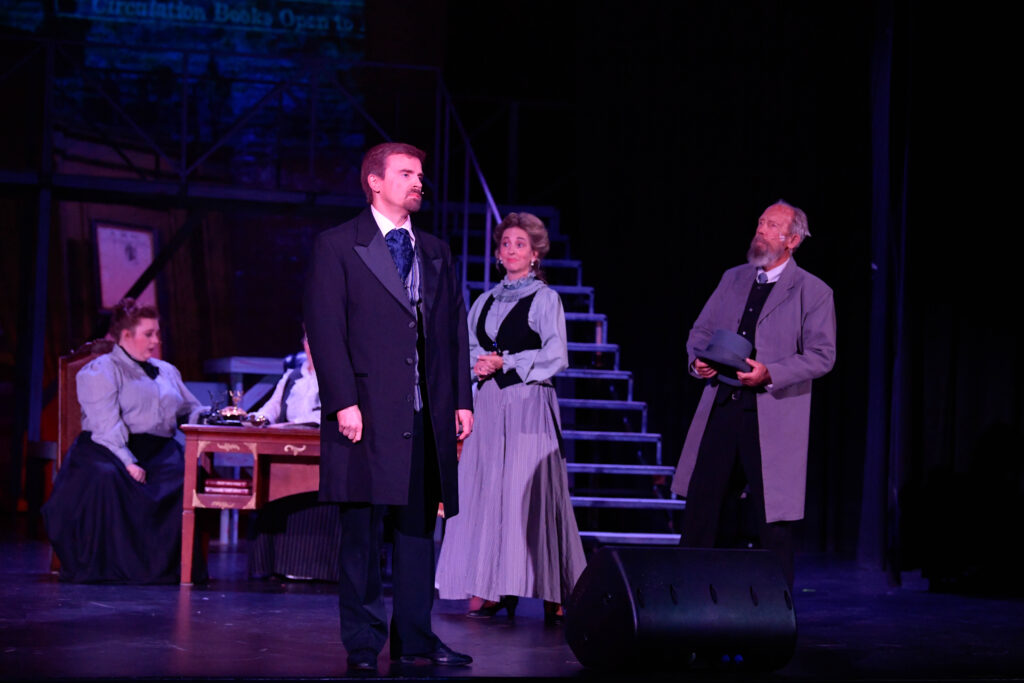 The image depicts a theatrical stage scene from the musical "Newsies." At the center, a man in a formal black coat and tie stands facing forward with a serious expression. To his left, a woman dressed in a long, high-collared, gray dress and blouse watches him attentively. To the far right, another man with a beard, wearing a long gray coat, holds a top hat. He also appears focused on the man in the center. In the background, a woman sits at a wooden desk, appearing engaged in work, with a typewriter or similar object on the desk. The setting includes steps leading upward on a dimly lit stage, with a projection of text faintly visible in the backdrop.