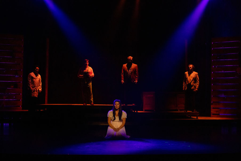 The image depicts a scene from a theatre production of "Bright Star." The stage is dimly lit, with blue and purple spotlights creating a dramatic atmosphere. In the foreground, a woman in a white dress kneels at the center of the stage, looking contemplatively into the distance. In the background, four men stand on an elevated platform. Each is dressed in brown or beige period attire, enhancing the historical setting of the scene. Wooden slatted walls flank the stage on both sides, adding to the rustic ambiance.