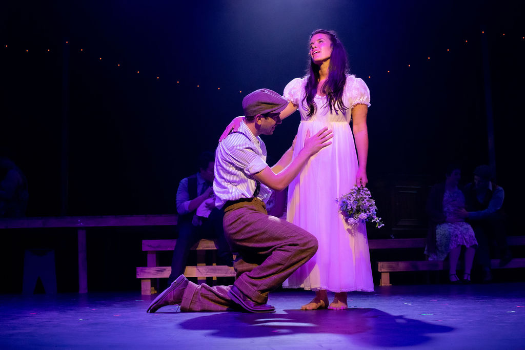 The image depicts a scene from a theater production, where a young man and woman are central on a stage. The man is kneeling on his right knee, wearing a flat cap, a light-colored striped shirt with rolled-up sleeves, suspenders, and brown trousers. He is looking up with a hand placed lovingly on the woman's waist. The woman stands barefoot, gazing distantly upwards, wearing a flowing white dress with short puffed sleeves. She holds a small bouquet of purple and white flowers in her right hand. Around them, dim lighting casts a soft, dramatic glow, with string lights visible in the background. Two seated figures are indistinctly visible further back on the stage, surrounded by minimalistic wooden set pieces.