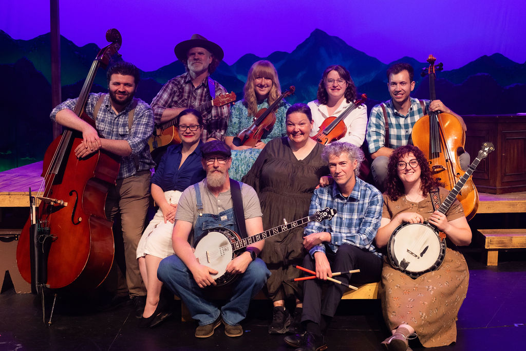 The image shows a group of ten people, arranged in two rows, posed on a stage set for a theater production. The background features a painted backdrop of dark blue and purple mountains against a vivid blue sky. The group is composed of eight men and women holding various string instruments, including a banjo, violin, double bass, guitar, and cello. They are dressed in casual, country-style clothing, with patterns like plaid and denim overalls. The person in the front row holds a drumstick alongside the banjo, and there are a variety of expressions, mostly smiling. The stage appears to be constructed from wooden platforms.