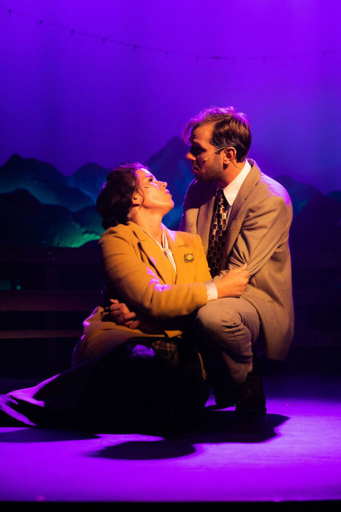 The image depicts a scene from a theatrical production of "Bright Star." Two actors are centrally positioned on stage under dramatic purple lighting. The female actor is kneeling, wearing a brown coat and looking up intently at the male actor. The male actor is crouching at her level, dressed in a light brown suit and patterned tie, with his face turned towards her. Both appear to be having an intense dialogue, their faces marked with subtle stage makeup. The background features a set resembling distant mountains, softly illuminated in green, with a string of small lights arcing above.