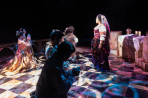 The image shows a stage scene from a theatrical production of "Hunchback of Notre Dame" by West Valley Arts. Several performers are dressed in period costumes. On the left, two women in golden, brocade gowns are kneeling with hands clasped in prayer. To their right, a man is also kneeling, holding a rosary. Centered is a woman standing, facing the kneeling individuals. She wears a richly colored dress with a patterned skirt and a burgundy bodice. Her hair is adorned with ribbons. In the background, stone-like pillars and an altar draped with a white cloth can be seen. Three lit candles are positioned on the altar. The floor displays a checkered pattern with colorful lighting casting various shapes.