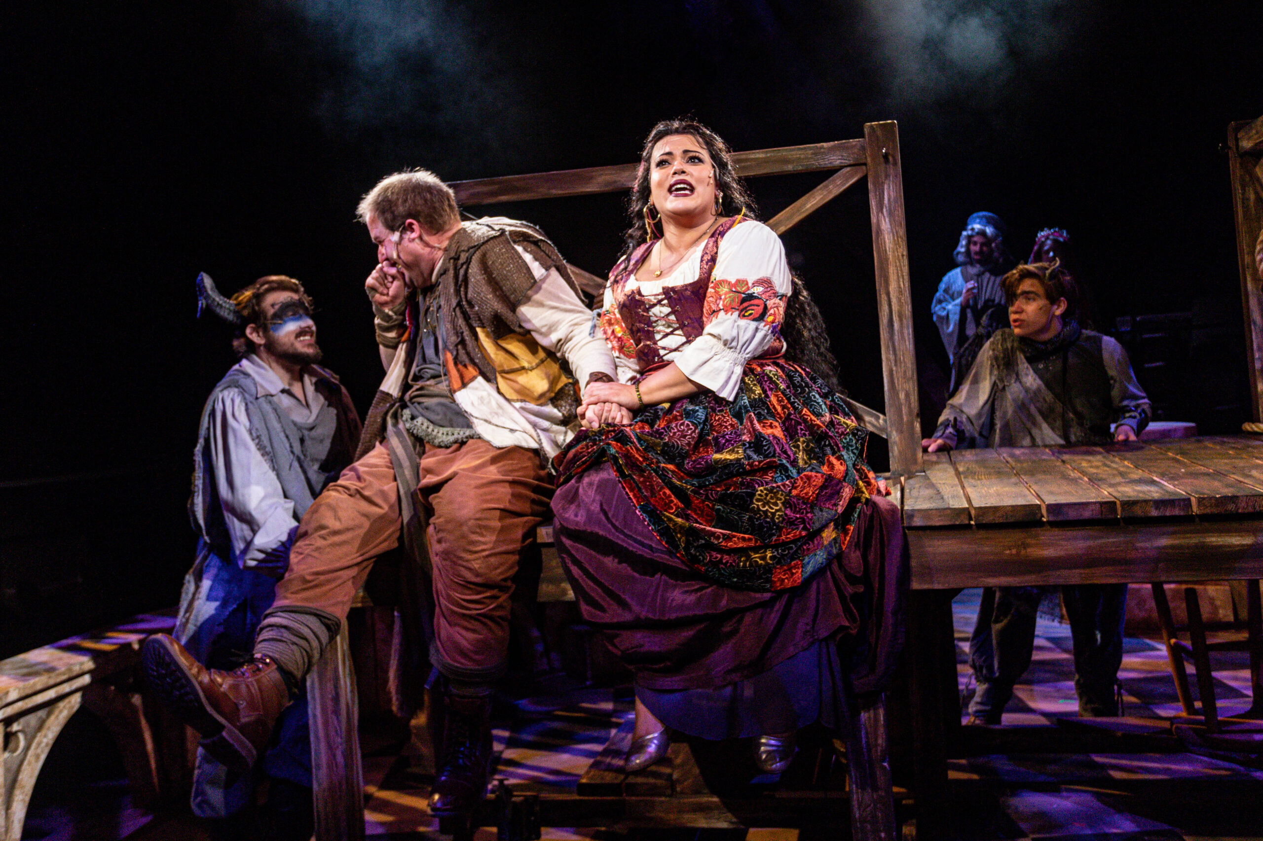 The image depicts a theatrical scene from the West Valley Arts production of "Hunchback of Notre Dame." Several actors are on stage, illuminated by dramatic lighting. In the foreground, a woman dressed in a patchwork, colorful skirt and white blouse is seated on a wooden structure, holding hands with a man wearing a distressed patchwork outfit with brown pants. To their left, another actor with a painted face and horns is seated on the edge of the structure, smiling. In the background, two other actors are visible, one with long hair and a headpiece, and another with short hair, listening intently. The scene is set against a dark backdrop, enhancing the theatrical mood.