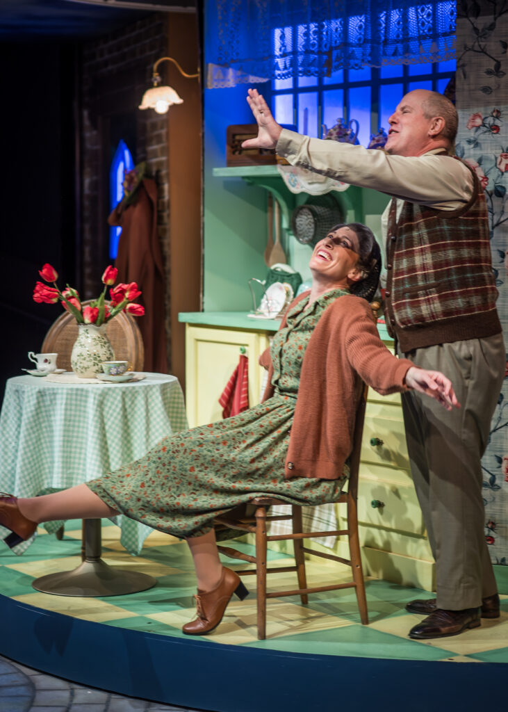 The image portrays a scene from the musical "Flowers for Mrs. Harris." A brightly lit kitchen setting serves as the backdrop. A woman, wearing a patterned green dress with a brown cardigan, is seated on a wooden chair, leaning back with a joyful expression. Her legs are crossed and lifted off the ground, wearing brown heeled shoes. A man stands beside her, dressed in a checkered vest and beige shirt, extending his arm outward as if gesturing or singing. A small round table to the left has a green checkered tablecloth, a vase with red tulips, and a set of teacups. The kitchen features yellow cabinets, mint green walls, and decorative plates. A window behind them is adorned with lace curtains and blue lighting, adding a lively and vibrant atmosphere. A hanging lamp and a coat rack with a brown coat are visible in the background.