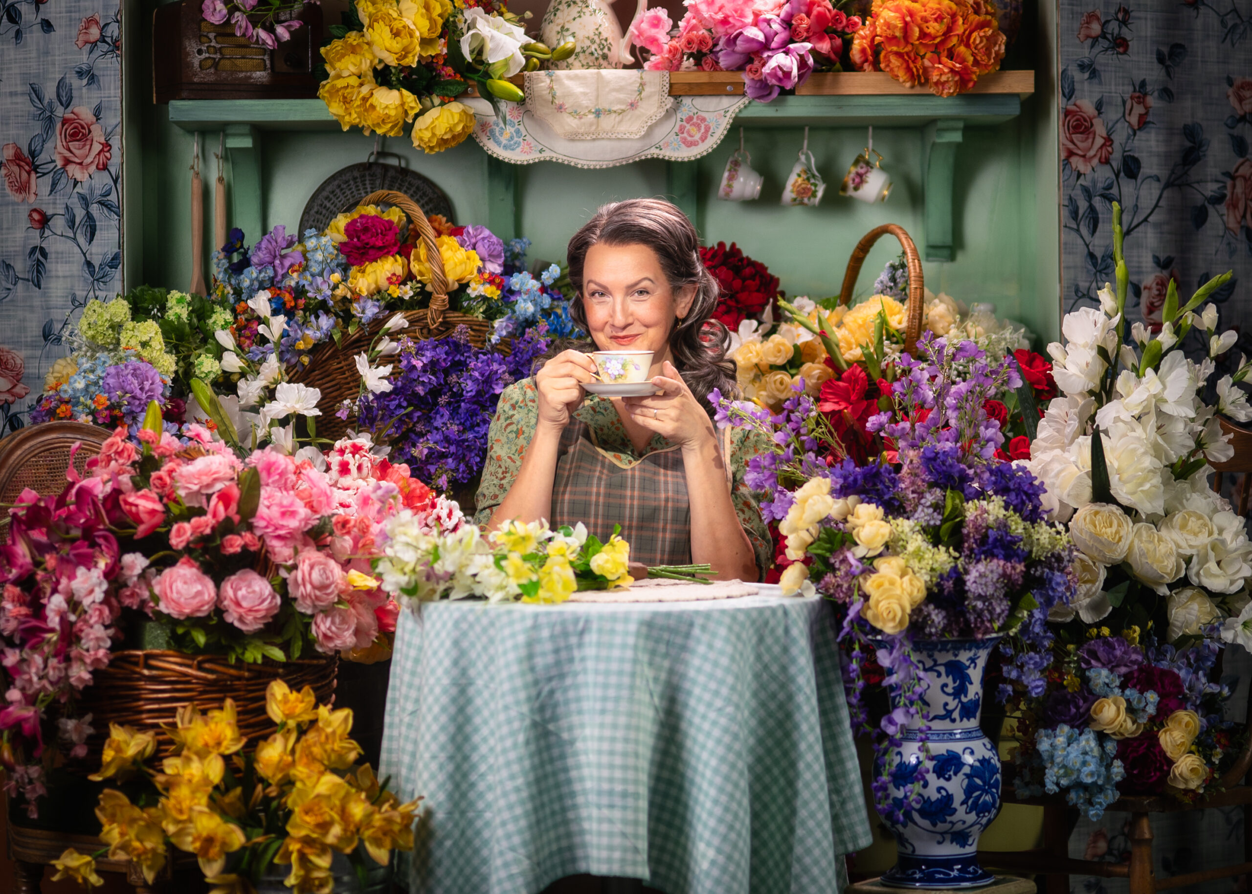 The image depicts a woman seated at a small, round table covered with a light blue and white checkered tablecloth. She is holding a teacup and saucer, smiling gently. The woman is surrounded by an abundance of colorful flowers in various shades of pink, yellow, red, purple, white, and blue, arranged in wicker baskets and vases. Behind her is a light green wall with hanging floral-patterned teacups and saucers. Floral arrangements are artistically displayed with a diverse mix of shapes and textures, creating a vivid, cheerful atmosphere.