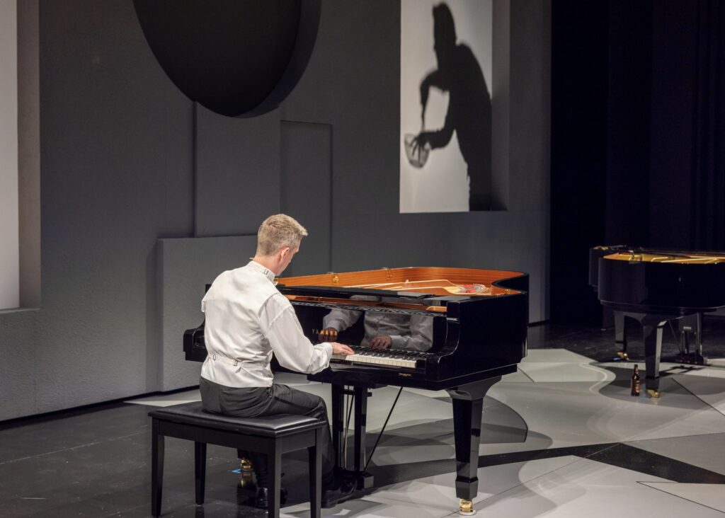 The image shows a person sitting at a black grand piano on a modern, minimalistic stage. The stage floor has geometric patterns in shades of gray. The pianist, wearing a white shirt, plays with his back to the camera. Reflections of his hands are visible on the glossy piano surface. To the right, another grand piano is partially visible. On the wall behind the pianist, a rectangular screen displays the shadow of a person stirring a whisk in a kitchen bowl, creating an artistic and slightly menacing silhouette.