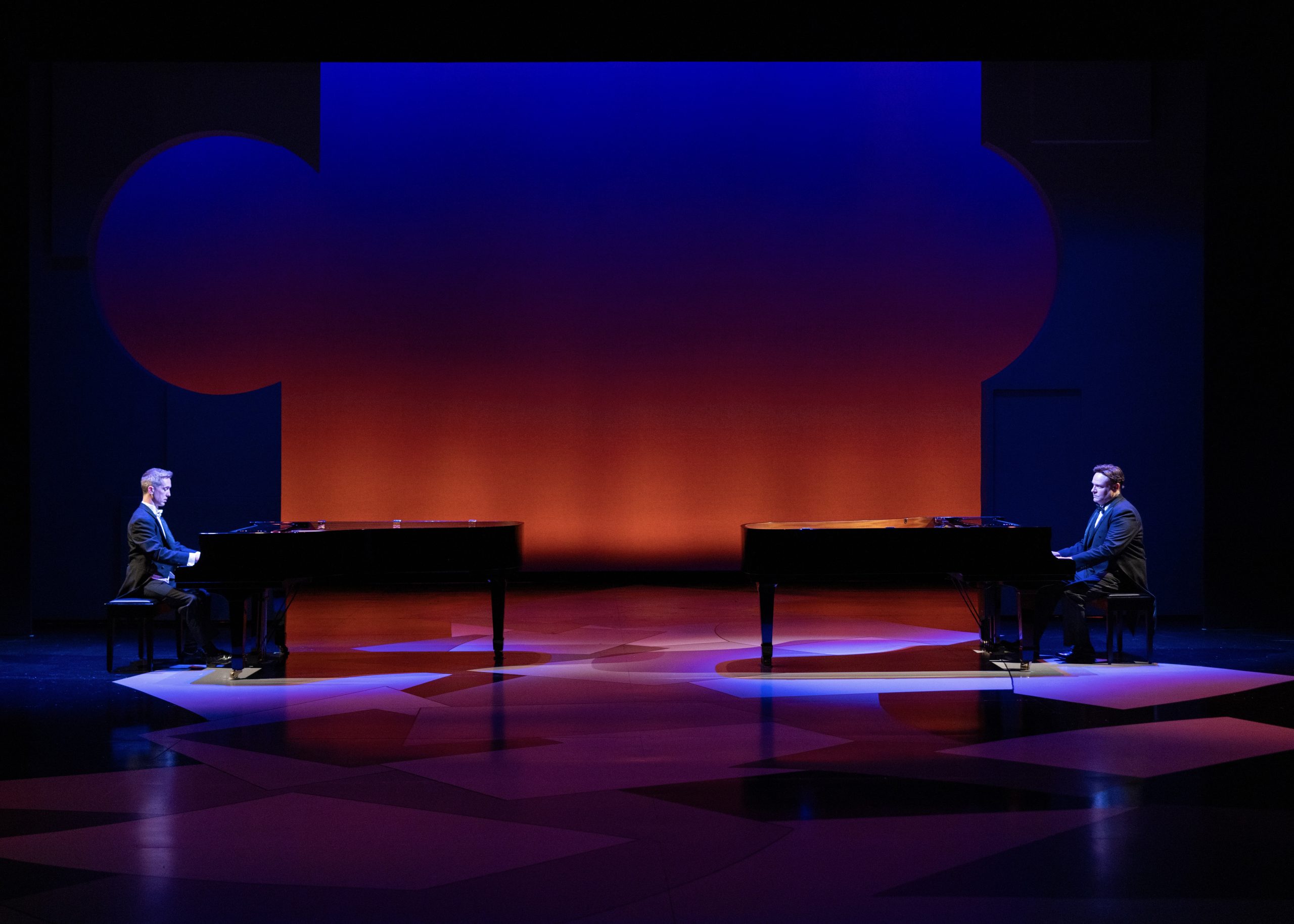 Matthew-McGloin-and-Richard-Todd-Adams-_-Credit_-BW-Productions-Res-374-scaled The image depicts a stage with two grand pianos positioned on opposite sides. Each piano has a person seated, facing each other. The stage is bathed in dramatic, colorful lighting with an orange backdrop that transitions to deep blue at the top. The floor features abstract geometric patterns illuminated with shades of blue and purple light. The individuals are dressed in formal black suits, each concentrating on their respective pianos. The overall atmosphere is serene and sophisticated, highlighting the concert-like setting.
