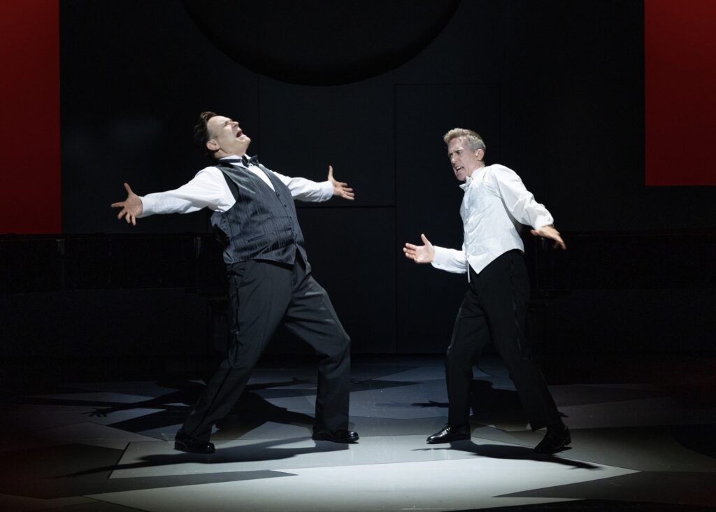 The image depicts two men on a dark stage performing with dramatic, open gestures. The man on the left is wearing a white shirt, black pants, black shoes, and a black and gray striped vest. He has his arms spread wide, leaning back slightly with an expressive facial expression. The man on the right is dressed in a white shirt with a white patterned vest, black pants, and black shoes. He also has his arms wide open and is leaning forward slightly, mirroring the expressive posture of the other performer. The background is dimly lit with shadowed geometric patterns on the floor and a large, subtle black circle on the rear wall. Hints of red are visible in the corners, adding contrast to the scene.