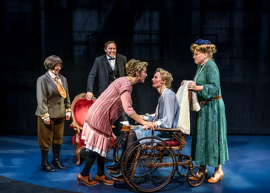 The image depicts a scene from the play "Silent Sky" on stage. Five actors in period costumes are positioned against a dark, blurred background. In the center, a woman in a light blue outfit is seated in a vintage wooden wheelchair. She has a gentle expression as another woman in a pink dress leans towards her, appearing to converse or console her. To their left, a man in a black suit with a bow tie stands behind a plush red armchair, smiling. A woman in a green dress with a blue hat stands to the right, holding a white shawl. A woman in a brown suit and knickers, along with knee-high black boots, stands on the far left. The stage floor is a smooth, deep blue, and the setting has a timeless, early 20th-century atmosphere.