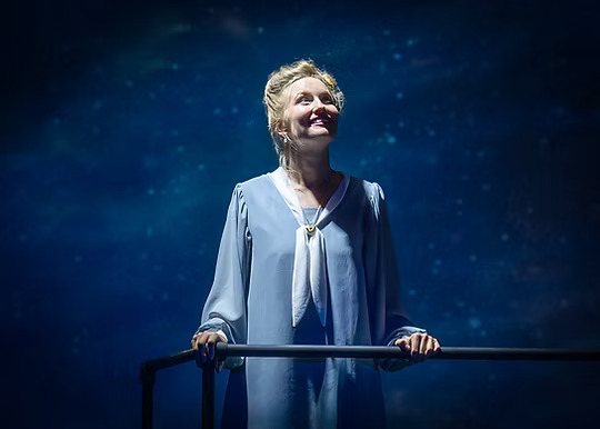 The image features a person standing on a stage, illuminated by soft, focused lighting against a backdrop resembling a starry night sky. They are wearing a light blue, long-sleeved dress with a white scarf-like collar tied at the front. Their hands rest gently on a dark metal railing, and they gaze upwards with a peaceful, thoughtful expression. The atmosphere conveys a sense of wonder and contemplation.