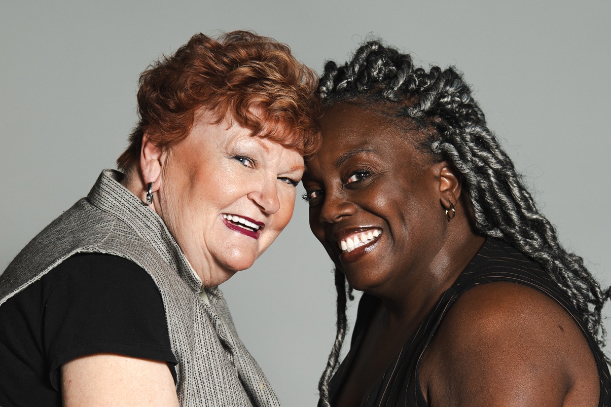 Two actors (a red haired, pale complected woman on the left, and a braided hair, dark completed woman on the right), putting their foreheads together for a publicity photo for Salt Lake Acting Company's "The Roommate".