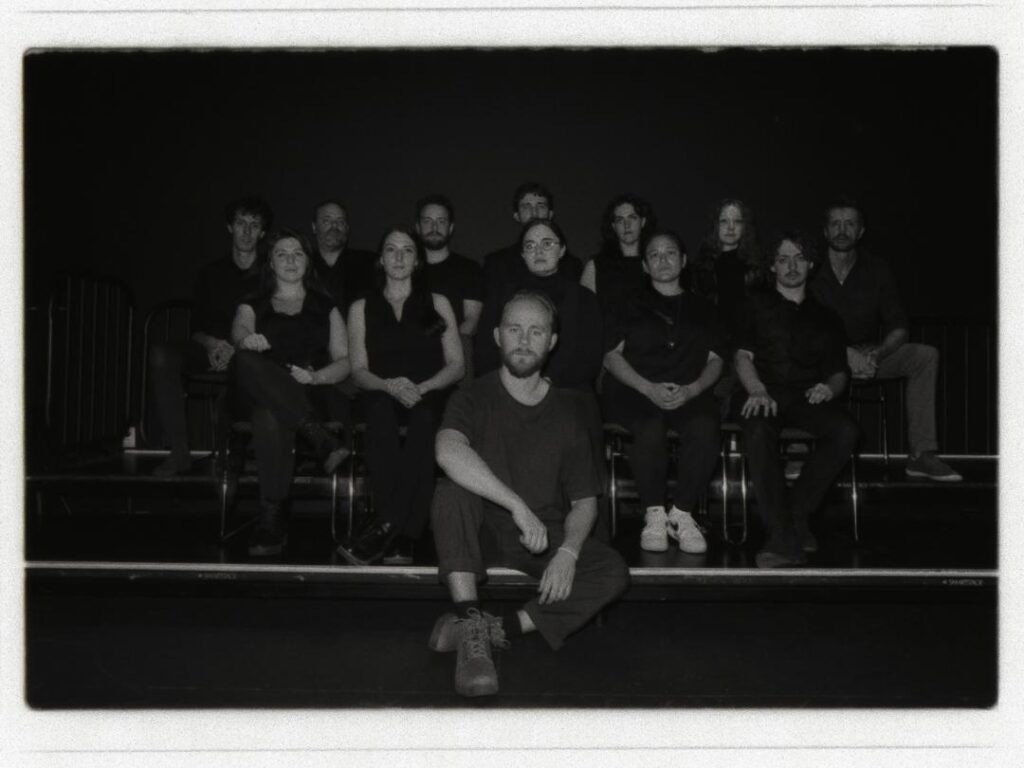 The black and white image depicts a group of thirteen individuals arranged in two rows on a tiered platform. The setting is dark, with the focus on the people. The front row consists of six individuals sitting side by side, all wearing dark clothing. In the center, one person is seated on the floor in front of the others, with legs crossed, wearing a dark shirt and boots. The second row consists of seven individuals who are also dressed in dark attire, seated closely together. The background is plain and unobtrusive, suggesting the photograph was taken in a studio or theater setting. The lighting is soft, creating subtle shadows and emphasizing the serious expressions of the group's members.