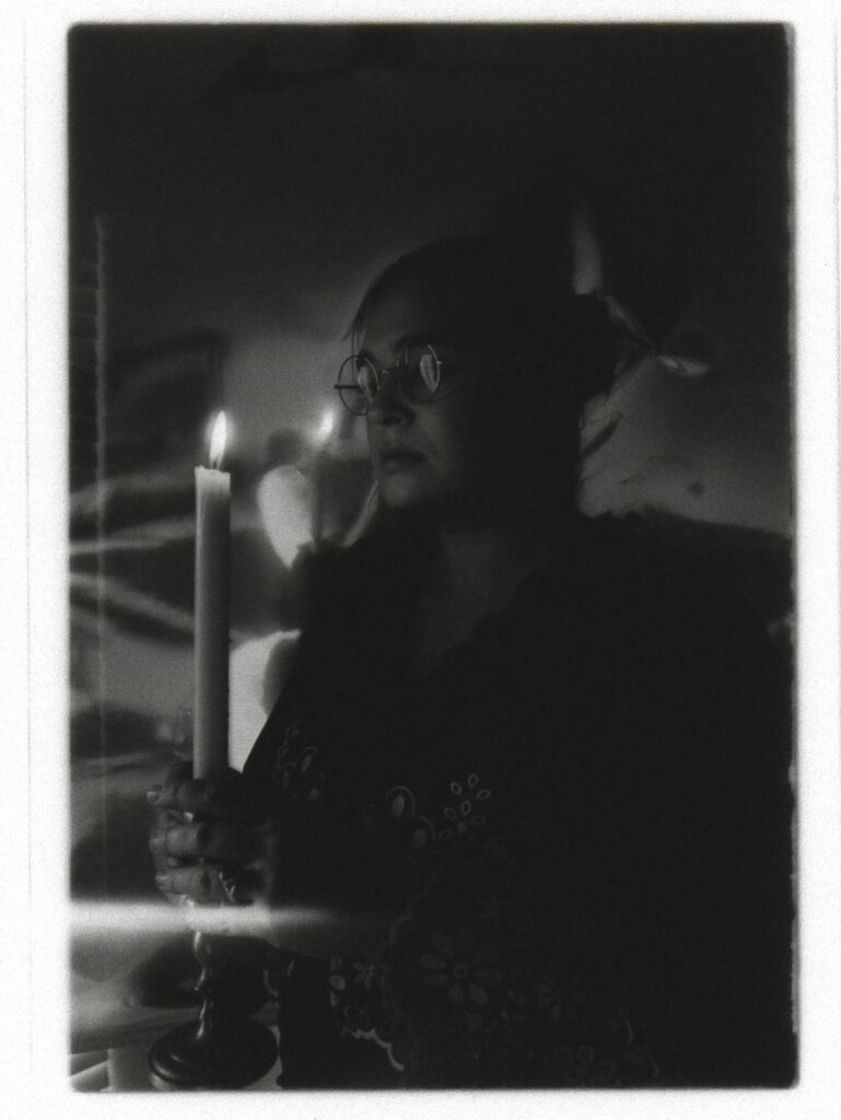 The black and white image depicts a scene with a person holding a lit candle. The individual is portrayed in partial profile, facing slightly to the side. They wear round glasses that catch reflections of the light, and their expression is contemplative. The person is dressed in a dark garment with intricate floral patterns. The soft focus and graininess of the image create a vintage or dramatic effect, enhancing the candle's warm glow, which is the sole light source, casting subtle shadows. The background is blurred and indiscernible, maintaining the focus on the subject and candle.