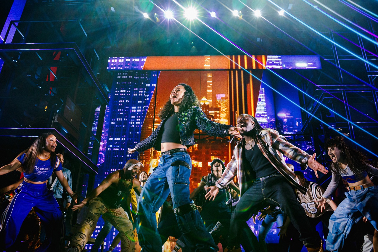 The image depicts a vibrant scene from a Broadway production. In the foreground, a diverse group of performers are energetically dancing with expressions of joy and enthusiasm. The central figure, with curly hair and a sparkly jacket, stands out with an open posture, surrounded by fellow dancers who are similarly animated. The background displays a dynamic cityscape projection with various skyscrapers lit up in vivid colors, creating a sense of depth and vibrancy. Bright stage lights beam from above, casting a lively illumination on the scene.