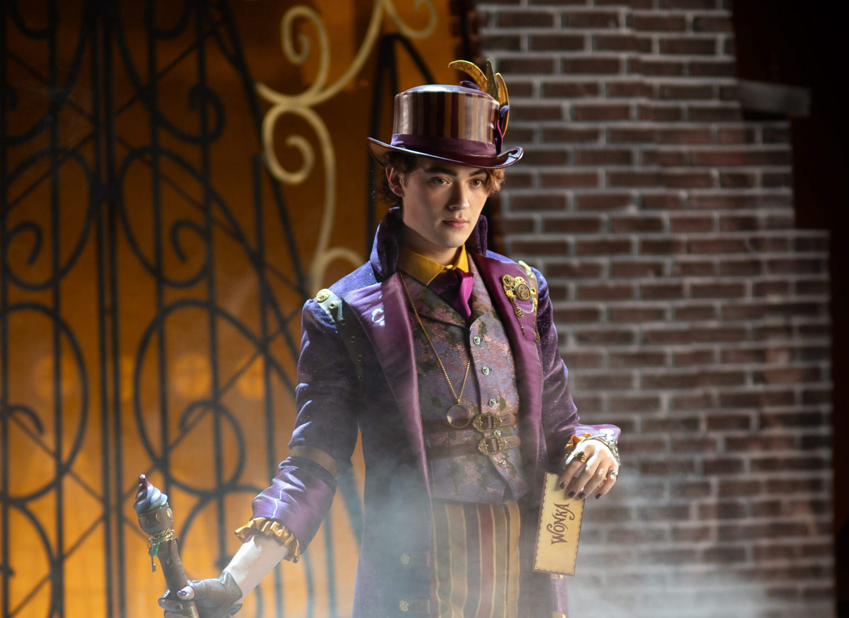 The image displays a theatrical scene from a production of "Charlie and the Chocolate Factory." A person is dressed in an elaborate outfit consisting of a purple coat adorned with gold buttons and embroidery. They wear matching purple trousers with vertical stripes in various warm hues. The ensemble includes a top hat with decorative feathers and a wide brim. The person stands confidently on a multicolored tiled floor, holding a polished cane in one hand and a golden ticket in the other. Behind them, a misty atmosphere envelops the scene, with a large, ornate iron gate partially visible against a backdrop of brick walls.