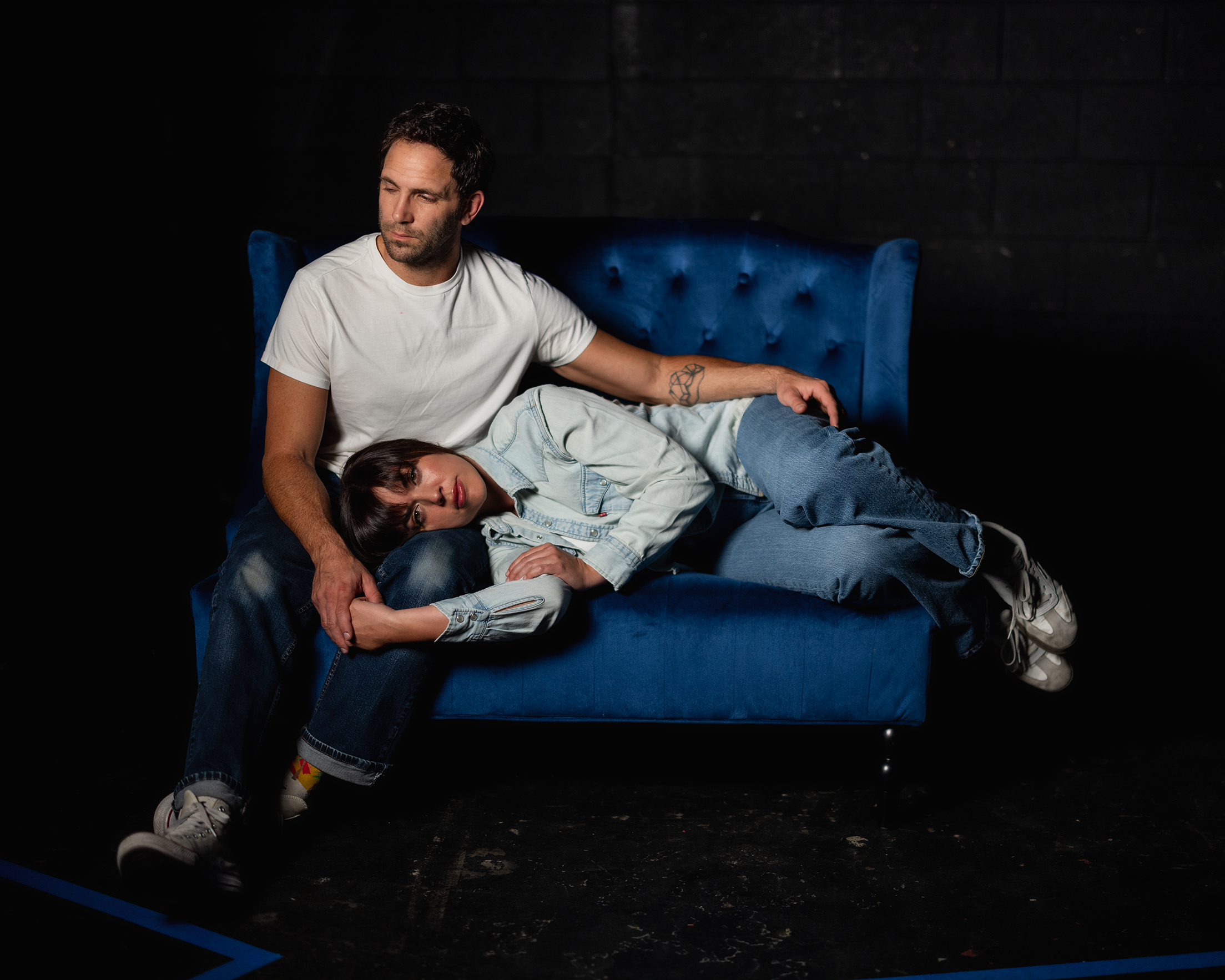 The image shows two people sitting on a bright blue, tufted-back sofa. The person on the left, wearing a white T-shirt and jeans, looks ahead with a thoughtful expression. Their arm is draped around the shoulders of a person resting their head on the other's lap. The person resting has long hair and is dressed in a light denim jacket and jeans, with their gaze directed towards the viewer. Both individuals wear casual sneakers. The background is dark, emphasizing the vivid color of the sofa.