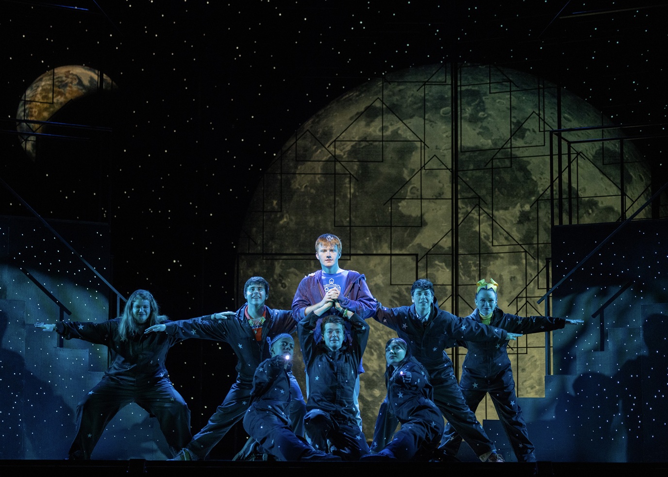 Carson Stewart and Company _ Credit_ BW Productions The image depicts a theatrical scene with a group of performers on stage. The central figure, standing slightly elevated, is framed against a large backdrop depicting a celestial body, possibly a stylized moon, with geometric patterns overlaying it. The stage is set in a space-themed ambiance, with a starry background and soft, cool lighting that highlights the performers. Surrounding the central figure are seven individuals, dressed in dark outfits adorned with star motifs, suggesting an outer space or cosmic theme. They are posed dynamically, with outstretched arms creating a sense of movement and unity. The setting is framed by stage elements resembling stairs on both sides, continuing the starry pattern.