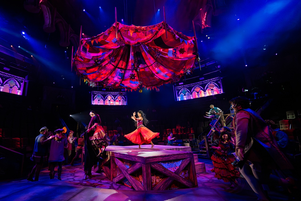 The image showcases a theatrical scene from HCT’s production of "The Hunchback of Notre Dame." The focal point is a large, elevated platform in the center of the stage, illuminated by vibrant lighting. A performer in a flowing red dress spins gracefully on the platform, creating a dynamic sense of motion. Above, a striking, large canopy of colorful, patchwork fabric hangs, adding a dramatic visual element to the stage. Around the platform, several other actors are engaged in animated expressions and gestures, clad in period costumes rich with color and detail. The surrounding stage area is enveloped in deep blue and purple lighting, highlighting the elaborate gothic architectural elements in the background with their stained-glass window designs. Spotlights and soft beams of light accentuate the theatrical atmosphere.