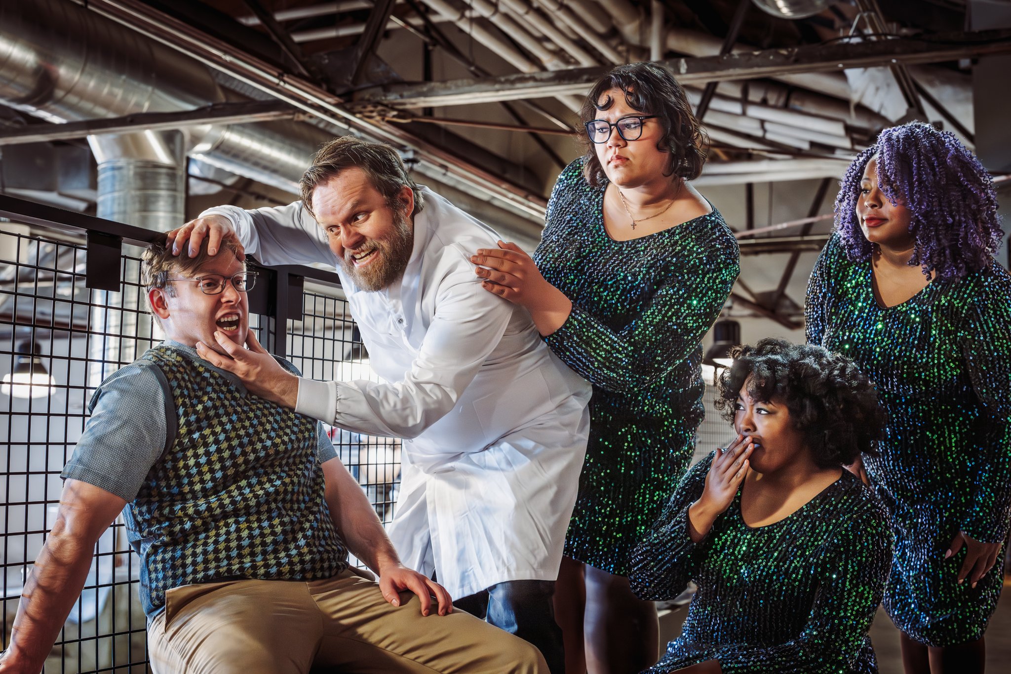 The image depicts a scene with five individuals in an industrial setting. The central figure, wearing a white lab coat and with a mischievous expression, is gripping the chin and head of a seated person who appears uneasy. This seated individual is wearing a patterned vest and glasses. Three other people, all in shimmering green dresses, are gathered around them. The one standing to the left is touching the central figure’s shoulder, while the person with purple hair watches to the right. The person at the bottom right, kneeling, has a hand over their mouth in surprise. The background consists of metal pipes and beams, characteristic of an industrial environment.