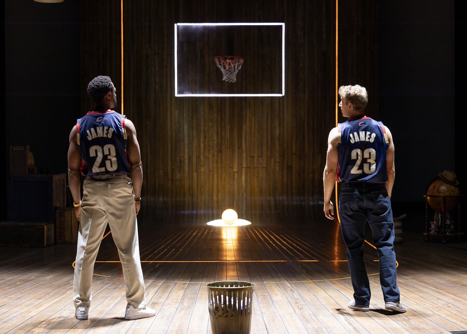 The image depicts a scene from Pioneer Theatre Company's play "King James." Two men, both wearing blue basketball jerseys with red and white trim featuring the name "James" and the number "23" on the back, stand on a stage. They are facing a wooden wall, which has a basketball hoop mounted on it, surrounded by a bright rectangular light. The floor is wooden and has a central line of light leading toward the hoop. A spotlight on the stage focuses on a basketball situated between the two characters. On the right side of the stage, there is an old-fashioned globe and a hat placed nearby. Both individuals stand with their hands by their sides, the one on the left dressed in light khaki pants and white sneakers, while the person on the right wears dark pants and shoes.