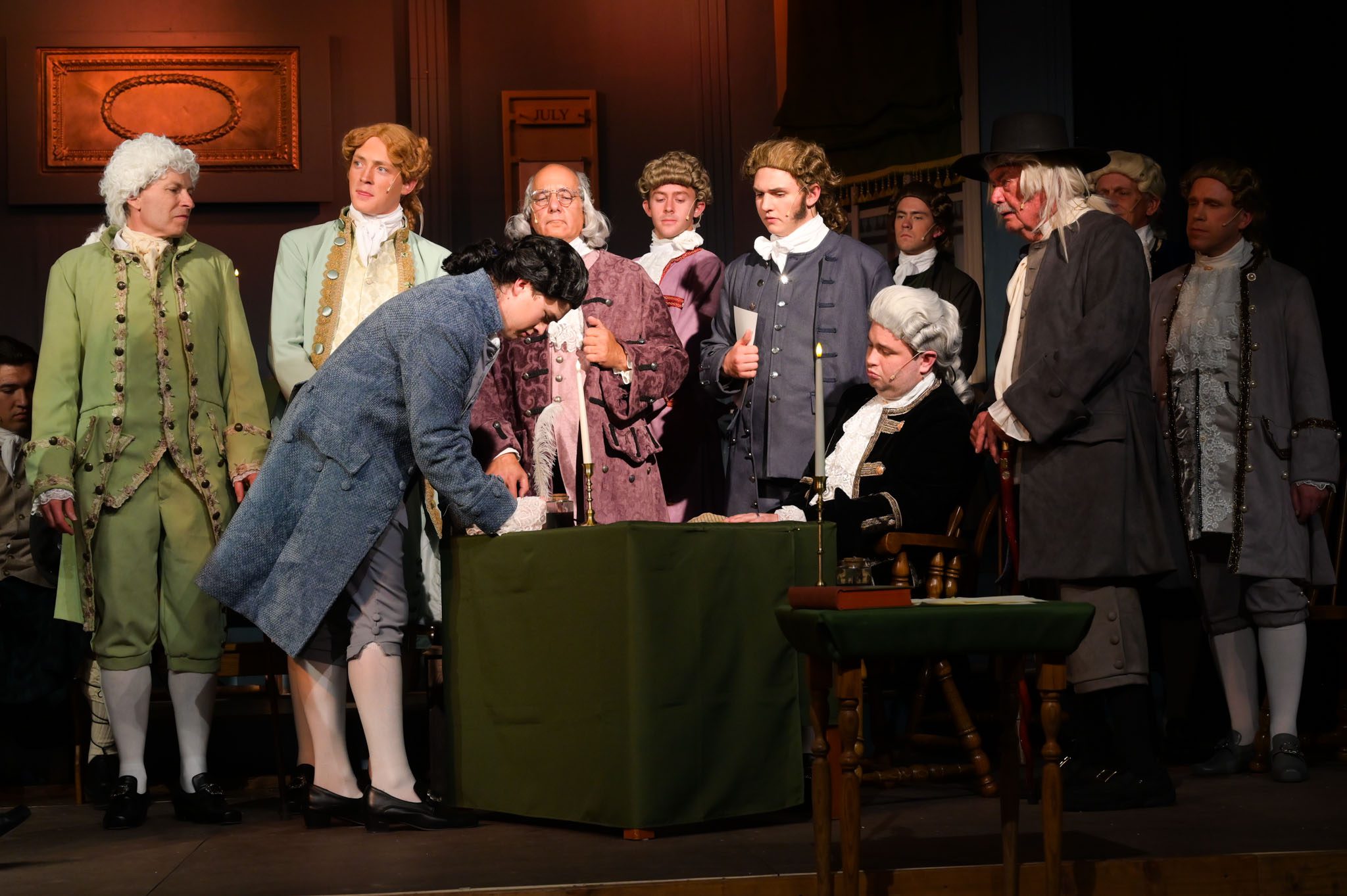 The image depicts a scene from the musical "1776," portraying the signing of the Declaration of Independence. The stage is set with a green table in the center, covered by a green cloth. A man in a blue coat is bent over, presumably signing a document, while several other historical figures, dressed in 18th-century attire with elaborate details, stand around him. To the left, two men in green and gray coats engage with the scene while a figure in a gray coat and a hat stands further back, observing. The characters have different hairstyles, most featuring wigs typical of the period, and exhibit a range of expressions reflecting concentration and seriousness. The background features a framed picture and a sign that reads "JULY," suggesting the historical context. The lighting creates a warm ambiance, focusing on the signing action.