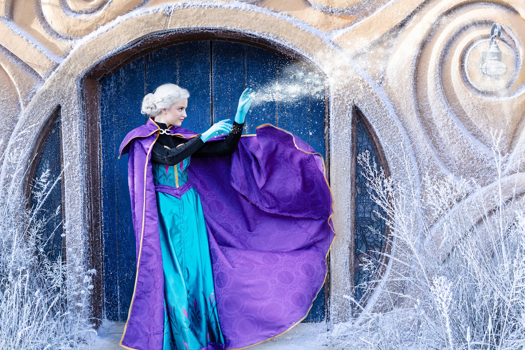 The image features a character [Elsa] dressed in a vibrant costume inspired by a winter theme. The character stands in front of a large, dark blue door framed by frost-covered, ornate, beige architecture with swirling patterns and decorative elements. The person is wearing a bright turquoise dress with purple accents and a detailed bodice. A rich purple cape flows dramatically as they gesture with gloved hands, from which sparkles of frost or snow seem to emanate. Around the entrance, there are icy branches enhancing the wintry atmosphere.